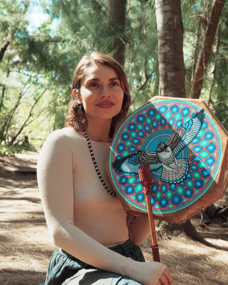 Brielle Águila sitting among pines, holding a painted frame drum