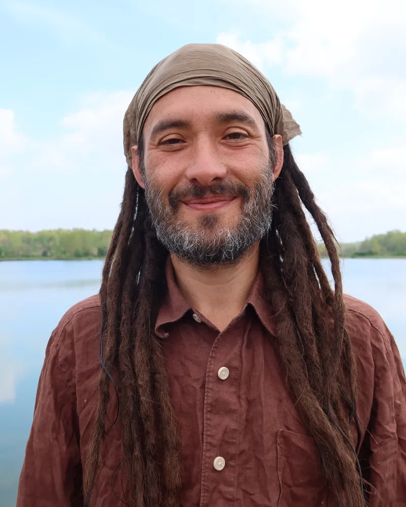 Eric smiling in a headscarf and a rust linen shirt, in front of a still lake