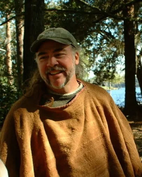 Johnny Dame smiling in a cap and a brown woven cloak, among pines beside a lake
